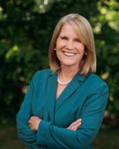 Woman with blondish straight mid-length hair, green suit jacket crossing arms and smiling